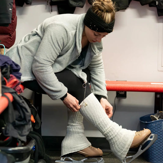 woman putting on figure skates in a locker room wearing a grey jacket and black pants and cream knit leg warmers 